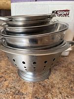 Stack of assorted metal bowls and colanders, showing multiple sizes and types including a colander with round holes and pedestal base.