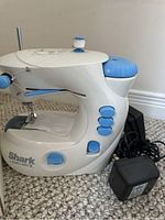 Side view of compact Shark Euro-Pro X sewing machine in white and blue with attached power cord and foot pedal visible on carpet.