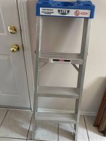 Front view of 4-step aluminum ladder with blue plastic tool tray top standing upright indoors.