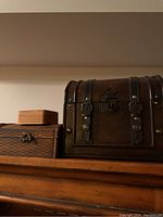 Photo showing three wooden storage boxes on top of a wooden surface: large dark brown chest-style box with black metal straps, smaller dark brown box with metal detailing, and a natural wood box with brass latch.