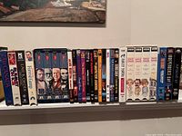 Photo of VHS tapes and DVDs arranged on white shelf, showing various titles: The Canadas, Law & Order, Home Gump, The Sopranos, The Matrix, etc.