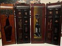 Front view of four wooden decorative wine boxes styled as antique books with dark brown faux leather spines and wooden bodies. Two boxes feature gold lettering 'The Pioneers 1829' and 'Gone With The Wind,' one has a painted wine bottle design.