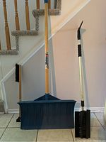 Three winter tools including a wide blue plastic shovel with wooden handle and two snow brushes with metal handles and black bristles.