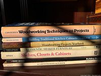 Photo showing spines of eight woodworking books stacked, titles readable reflecting woodworking techniques, cabinetry, furniture, and project planning topics.
