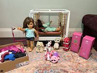 Overall lot showing two American Girl dolls, white canopy bed with pink beaded curtains, two pink lockers, luggage pieces, several stuffed animals, and a box of doll clothing.