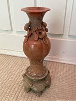 Full view of the antique reddish-brown carved jade vase with floral motifs on neck and a decorative carved stand, placed on carpet in front of white wall paneling.