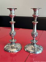 Pair of antique silver plated candlesticks displayed on a red surface with visible grape and leaf motif detailing on the stems and bases.