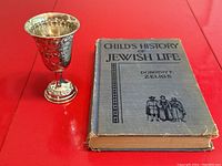 Photo showing the Kiddush cup next to the book on a red surface with clear view of the book's title and general condition.