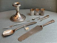 Photo showing entire lot including silver candlestick, pedestal dish, three silver shot glasses, two forks, two spoons, serving tongs, and cake lifter.