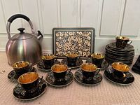 Full display showing 8 black and gold marble-patterned teacups with matching saucers, 10 side plates, a large stainless steel kettle, and black napkins along with floral patterned placemats.