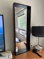 Full view of the large contemporary black mirror standing upright on a surface next to a lamp and display items, showing its reflective surface and deep frame.