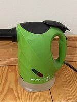 Front side view of green H2O SteamFX Pro handheld steam cleaner on wooden surface, showing handle and indicator lights.