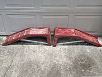 Pair of red metal car ramps laid out side by side on concrete surface in front of garage door.