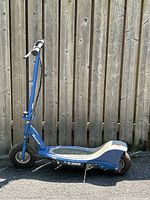 Full side view of blue Razor E300 electric scooter with foot deck, handlebar with white grips, and rear kickstand visible. Scooter has thick black tires and blue frame.