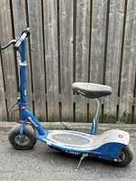 Side view of blue Razor E300 electric scooter with black seat, standing on pavement in front of wooden fence.