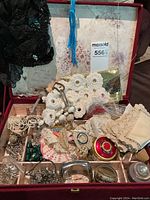 Top-down view of vintage costume jewelry and crocheted handbags in a compartmentalized box. Includes brooches, necklaces, bracelets, earrings, lace, and small unique items like a round red trinket box.