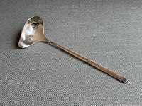 Full view of the sterling silver Georg Jensen serving ladle lying diagonally on textured gray fabric, showing entire ladle from handle to bowl.