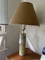 Vintage onyx table lamp standing on a wood surface next to a window showing natural and greenish striations on the cylindrical onyx base with a beige fabric shade decorated with hanging beads along the bottom edge.