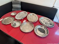 All eight silver plated serving trays displayed on a red table, showing various sizes and shapes including oval and round trays with decorative edging and embossing.