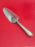 Close-up of the cake cutter/lifter showing the comb-style cutting blade and sterling silver handle with decorative engraving on red background