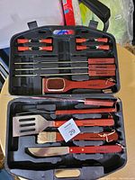 Photo of BBQ tool set inside black plastic carrying case showing skewers, forks, spatula, basting brush, and knives with wooden handles.