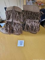 Pair of taupe suede fringe boots with multiple layers of fringe and decorative stitching at the top, shown on a yellow table.