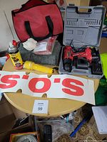 Photo of car safety kit items including red carrying bag, yellow flashlight, grey cloth, Fix a Flat spray can, white rags, first aid kit, and red/black cordless drill in plastic case with charger.