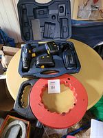Photo showing cordless drill, battery, charger in black carrying case, and a red fish tape wire puller on a round wood table.