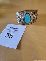 Photo of Tibetan silver cuff bracelet with central turquoise stone on wooden surface next to auction lot card