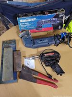 Photo showing blue Mastercraft detail sander with power cord, original packaging box, black soldering tool with cord, two sharpening stones, and red-handled pliers on a table.