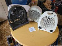 Photo of three units on a round wooden table: one black table fan and two white combination fan/heater units, showing front views and controls.