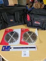 Two circular saw blades in original packaging, one hacksaw blade in packaging, and two black tool bags on a table.