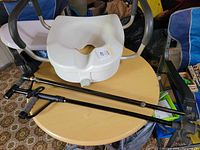Photo shows a white raised toilet seat with arm handles placed on a round wooden table, along with two black adjustable canes. One cane has a standard rubber tip and the other cane features a rubber tip plus a built-in light.