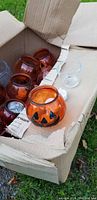Cardboard box with multiple small orange translucent glass pumpkin holders, some decorated with jack-o'-lantern faces, and a few clear glass tea light holders visible inside and on edge of box.
