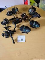 Photo showing eight assorted fishing reels arranged on a round wooden table with a MaxSold lot card number 28.