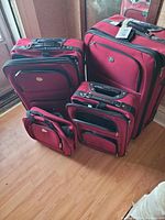 Four-piece red luggage set on wooden floor near window, showing all four pieces in good condition.