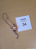 Photo of a silver-tone necklace with a round Poke Ball pendant on a wooden surface next to a numbered auction slip.