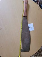 Full view of the antique Crocodile brand sugar cane machete laid flat on a light round wooden table with yellow measuring tape alongside showing 22 inches length.