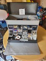 Photo showing open metal tool box with multiple clear compartments holding various small hardware and electrical parts