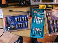 Overhead photo of a blue blister pack containing 10 drill bits, a blue card packaging 10-piece Dremel polishing and grinding stone set, and a brown blister pack of drill bits.