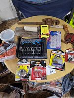 Image showing assorted hand tools and hardware laid out on round wooden table including rusty pipe wrench, hole saws, locks, multimeter, tape measure, mallet, screwdrivers in case, wire stripper, wires, and various small parts.