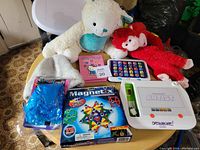 Photo shows a table laid out with various children's toys including a Magnetix magnetic building set box, multiple stuffed animals, two electronic learning tablets, and a white electronic artist toy with colorful controls.