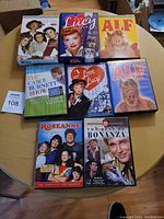 Eight DVD sets on a wooden table, displaying box covers for classic TV shows including I Love Lucy, Alf, Carol Burnett, Roseanne, and Bonanza.
