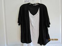 Front view of black shrug over white cami hung on hanger against white background.