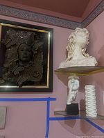 Framed wall art sculpture with woman's face surrounded by butterflies and textured elements, hung on a pink wall with other sculptures on glass shelves.