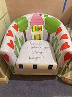 Front view of colorful child's chair showing painted houses, trees, hearts, and white dots on a lavender background, with the phrase 'home is where your story begins' written on the seat cushion.
