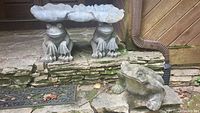 Three concrete garden frog statues placed on outdoor stone steps near wooden siding and a downspout.