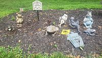 Wide view of the lot showing multiple frog-themed garden ornaments including statuettes and metal signs arranged on mulch.