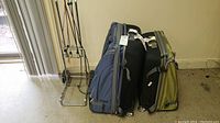 Three soft-sided suitcases in blue, black, and green colors stacked against a wall with a metal luggage cart next to them.
