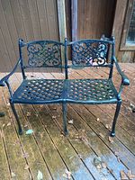 Full front view of black cast metal patio bench on a wooden deck, showing lattice seat and decorative back panels.